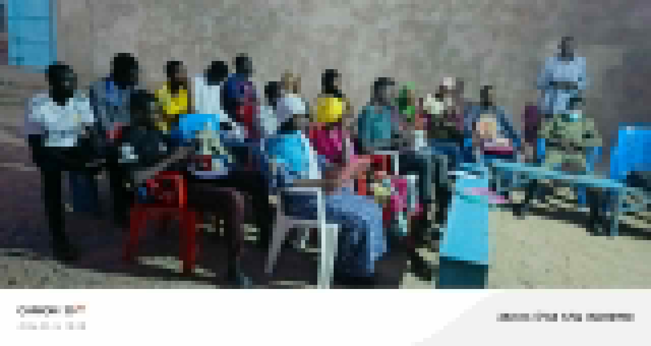 Students seated in a long row outdoors at the Abeche Center, in front of a tall stucco wall, men in lighter shirts on the left, women in bright wraps and dresses on the right.