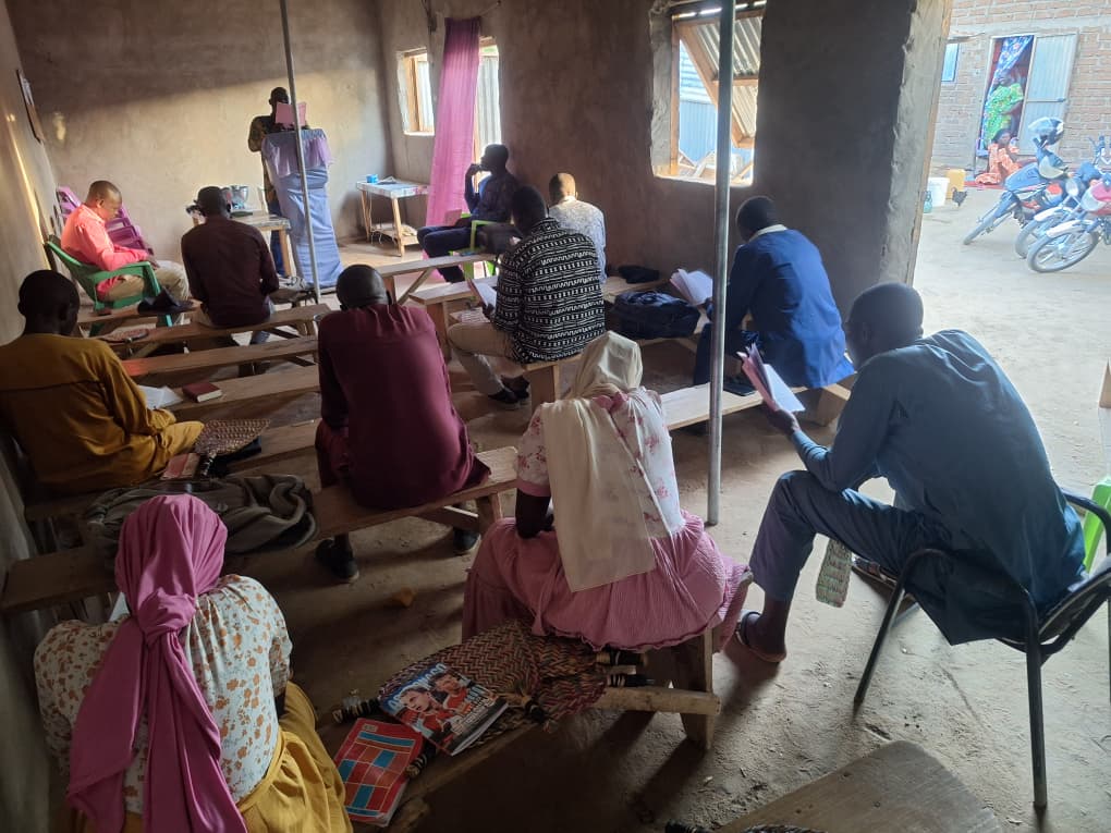 Men and women gathered for Portable Bible School in Amdourmile, West Chad — wooden benches in a brick-walled room, a motorbike resting at the open doorway.