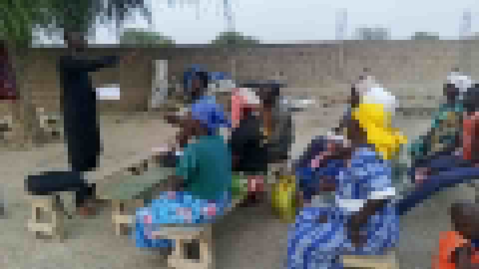 Outdoor gathering at the Amdourmile center. A teacher stands at left, students seated on low wooden benches under open sky, women in bright wraps holding their books.