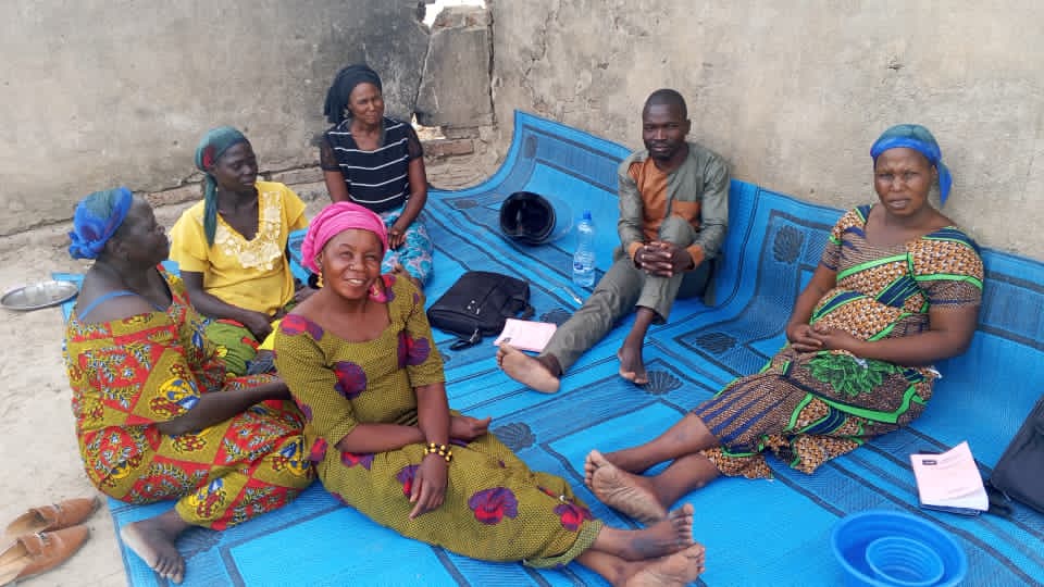 A smaller study group at Amdourmile seated on a blue patterned mat against a concrete wall. Women in colorful wraps and a man in an olive shirt, paperwork in their laps.