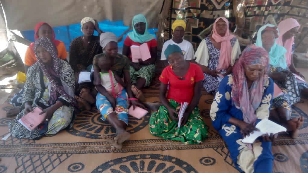 A row of women in red, blue, pink, and patterned wraps seated on a tarp under a blue shelter, pink Bible booklets open across their laps.