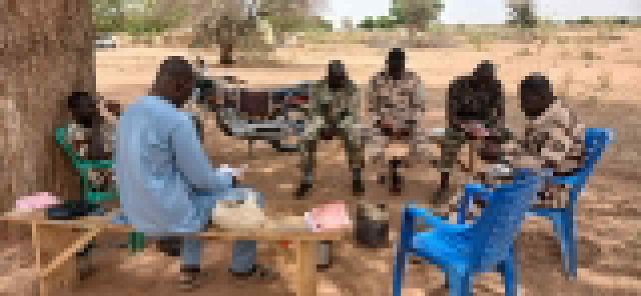 A teacher in a light blue shirt sits across from a small group of soldiers in camouflage, benches and chairs arranged in a circle under a tree.