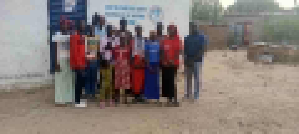 A group photo of Nord Ou'est students standing together in front of a small building, women in red and patterned dresses in the front row.