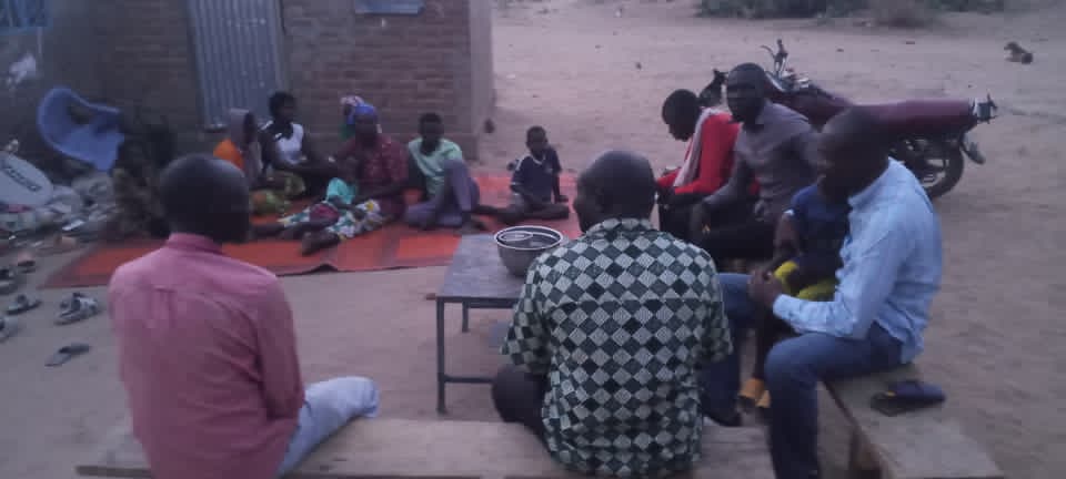 Outdoor evening gathering at Nord Ou'est. A small group of men seated on benches around a low wooden table, women and children on mats in the background, a motorbike at the edge of the scene.