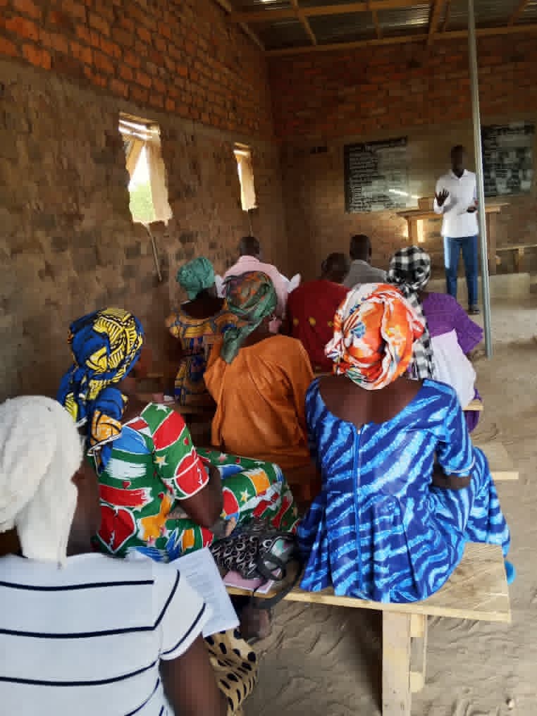 Women in vivid headwraps and dresses seated on benches inside a brick walled room at Selah Manga, a teacher in a white shirt standing at the front.